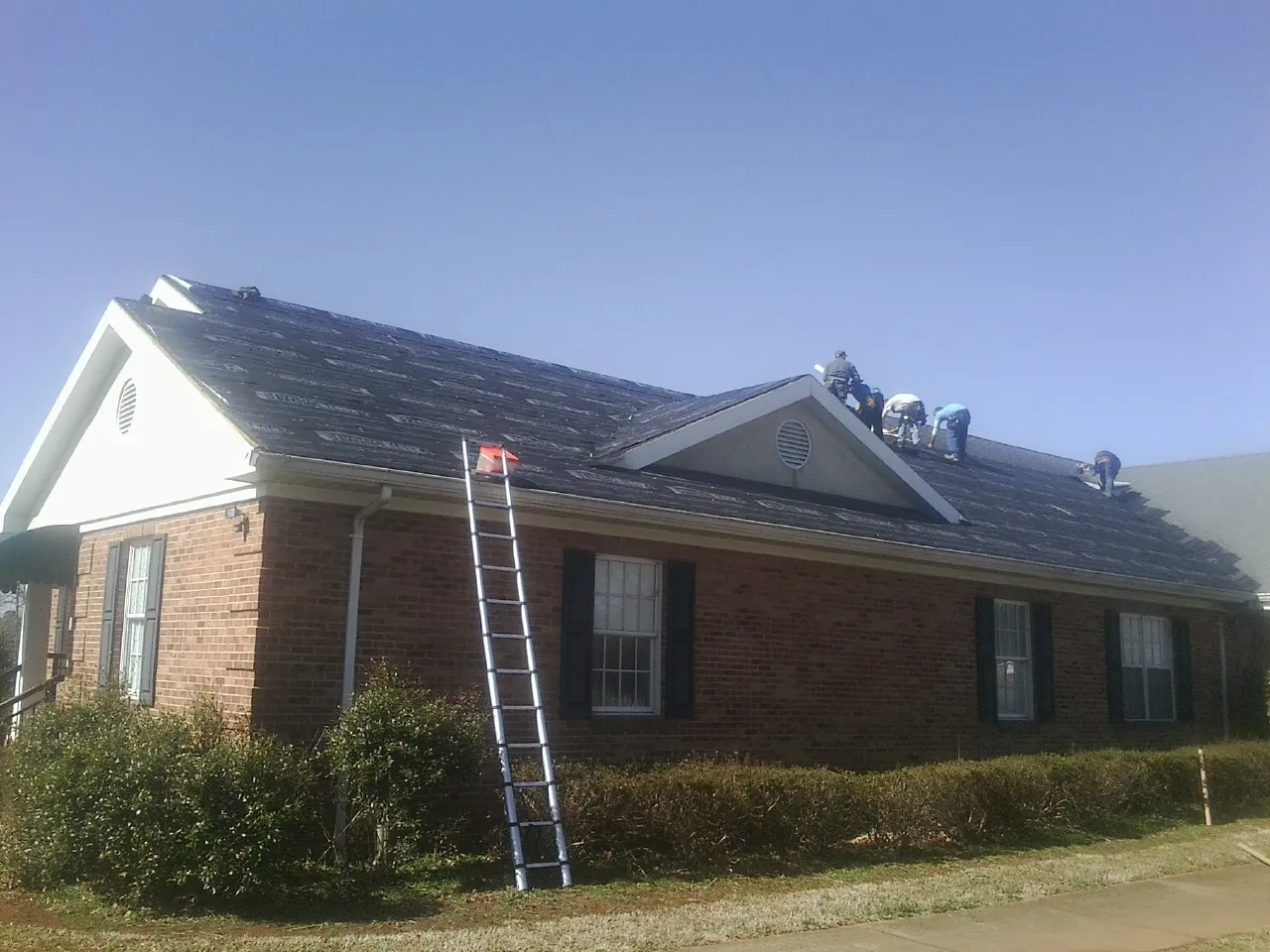 Crew tearing shingles off a brick ranch roof with a ladder set — Metal Roof Repair work by our crew in Howard
